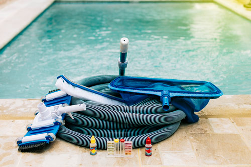 Reinigungszubehör wie Bodensauger, Bürsten, Kescher und Wassertest-Set vor einem Pool, um Ablagerungen am Boden zu entfernen.