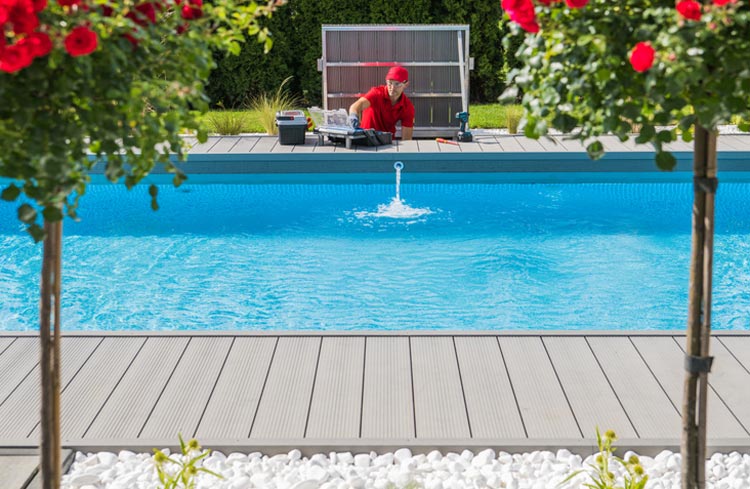 Ein Techniker in roter Arbeitskleidung repariert einen Pool im Garten. Im Vordergrund befinden sich dekorative Blumen und weiße Kieselsteine, die das moderne Design des Pools betonen.